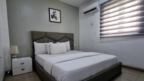 A bedroom with a bed, a nightstand, a lamp, a framed picture on the wall, and a window with blinds. - Quintessential Hotel Port Harcourt (Port Harcourt)