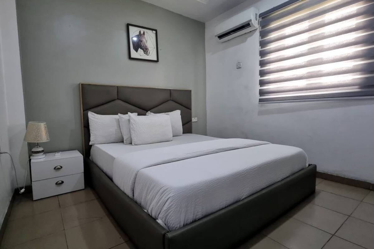 A bedroom with a bed, a nightstand, a lamp, a framed picture on the wall, and a window with blinds.
