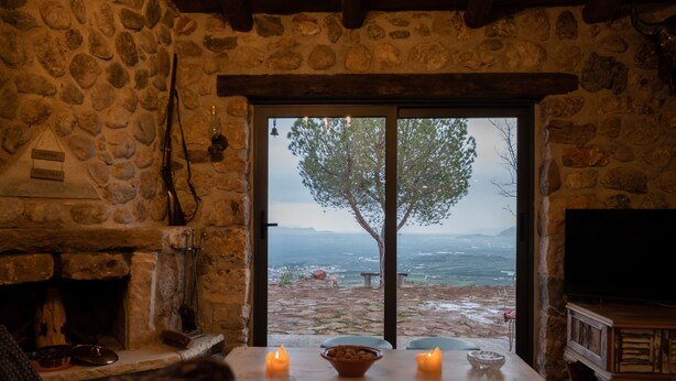 Habitació de parets de pedra amb una llar de foc, una taula amb espelmes, i una vista d'un arbre i un paisatge a través d'una porta de vidre.