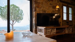 Paret de pedra amb un televisor sobre un suport de fusta, una finestra que mostra un arbre i el mar, i una taula amb un bol de vidre i un objecte groc.