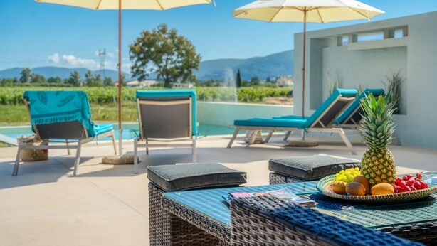 Zona de piscina con sillones de descanso, sombrillas, una mesa con frutas y vistas a las montañas.