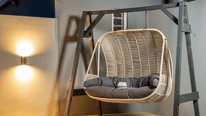 Una silla colgante de mimbre con cojín, suspendida por una estructura metálica, frente a una pared con dos luces redondas.
