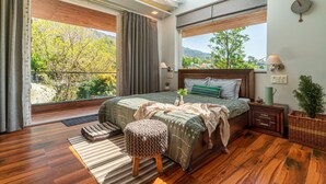 A bedroom with a large window, wooden flooring, a bed with green bedding, a bedside table with a lamp, and a small plant. - Celestial Rhapsody (Nainital)