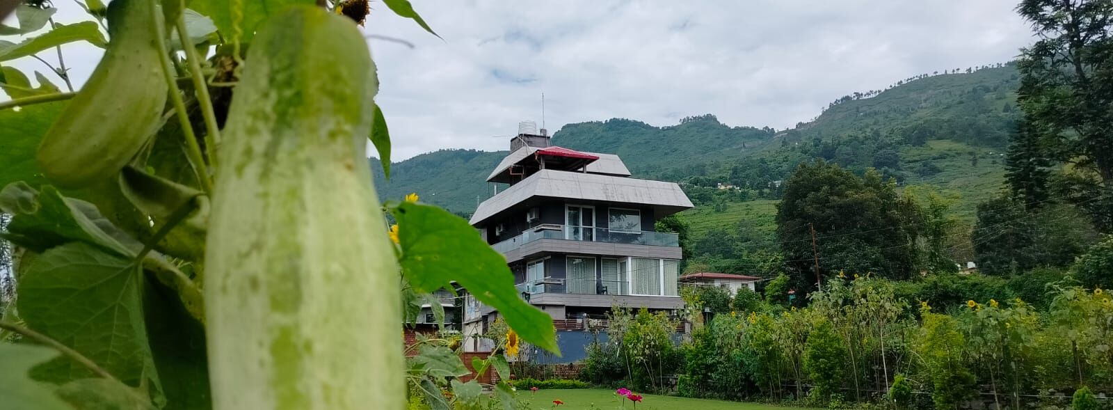 A modern two-story building with a flat roof and a balcony, surrounded by lush greenery and hills in the background.