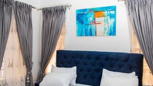 A bedroom with a large bed, grey curtains, and a blue headboard. - Delight Apartments Oniru VI (Lagos)
