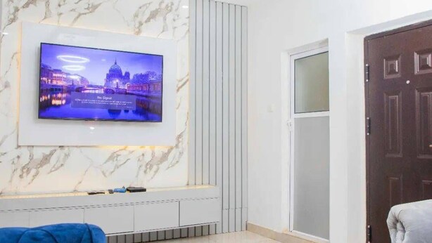 A modern living room with a flat-screen TV mounted on a marble wall, a grey sofa, and a coffee table with decorative items. - Delight Apartments Oniru VI (Lagos)
