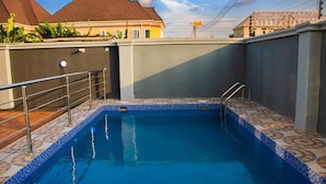 Ein Schwimmbecken mit gefliesten Rändern und Metallgeländern, umgeben von einer niedrigen Mauer und einem Gebäude mit einem Ziegeldach im Hintergrund.