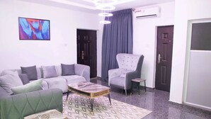 Ein modernes Wohnzimmer mit einem grauen Sofa, einem passenden Sessel und einem Couchtisch. An der Wand hängt ein Gemälde und eine Deckenleuchte ist angebracht.