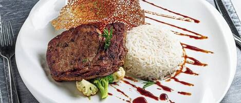A plate with a steak, rice, and vegetables, garnished with sauce, on a dark table.