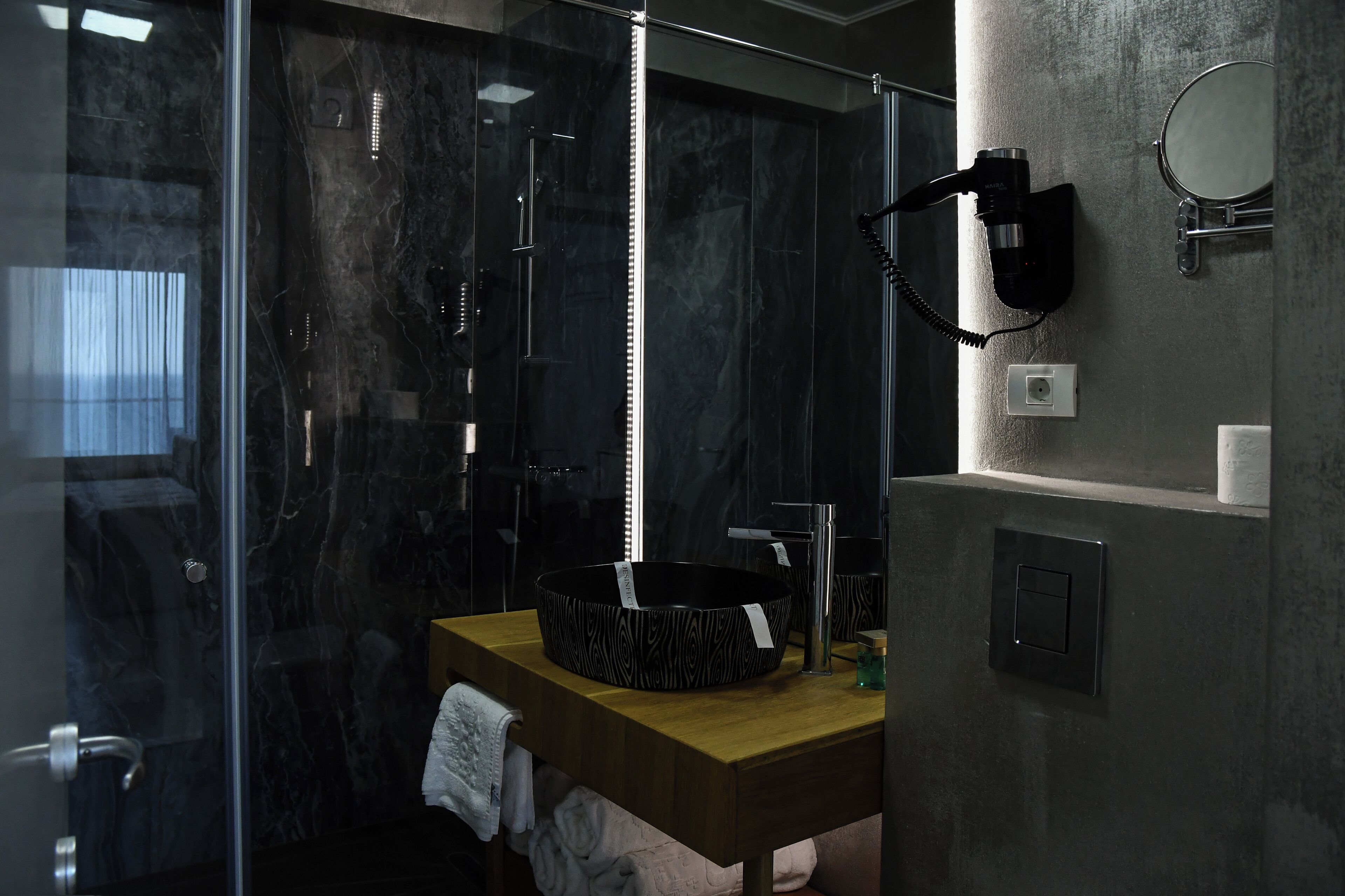 A modern bathroom with a glass-enclosed shower, a wooden vanity with a black patterned sink, and a hairdryer mounted on the wall.