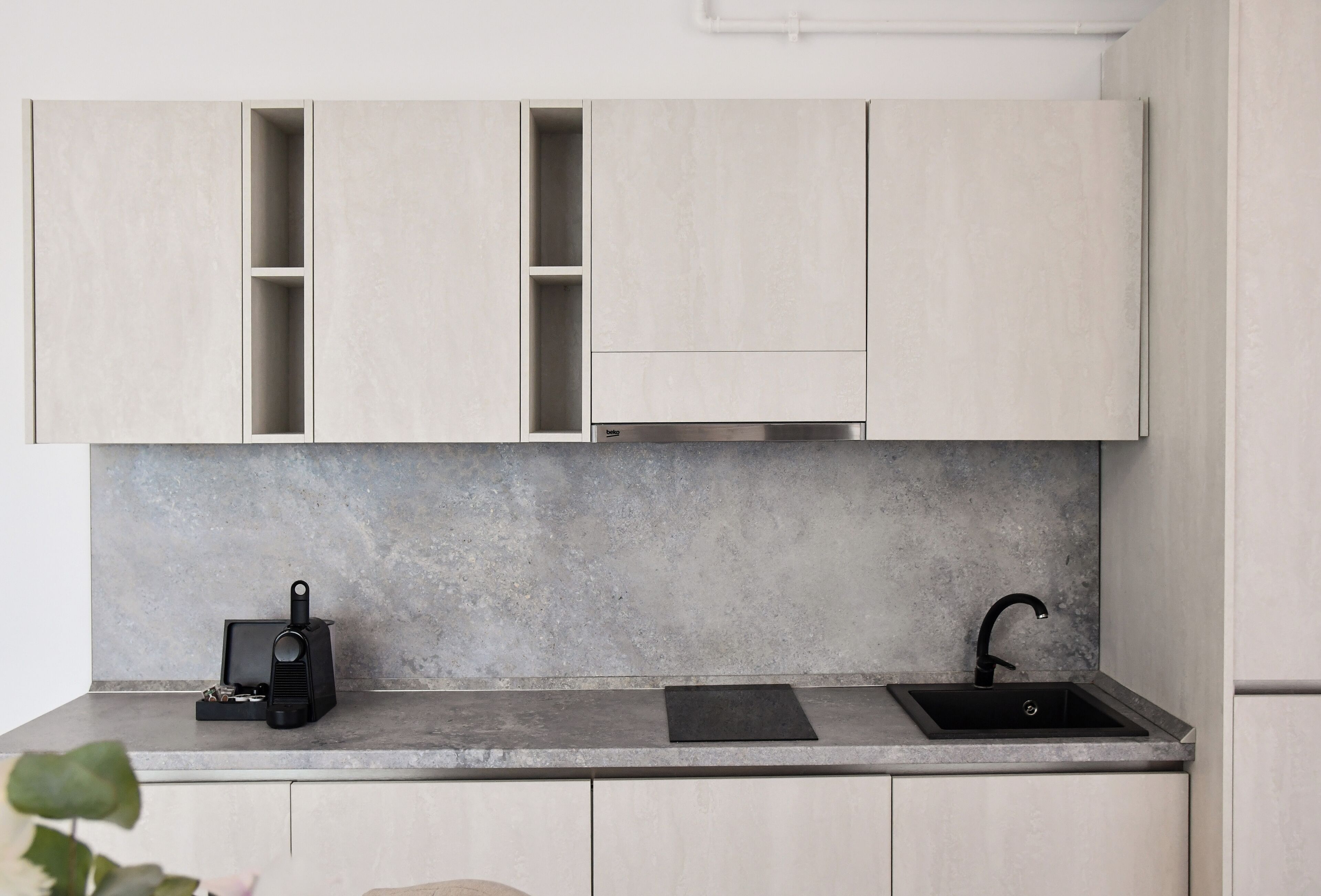 Una cocina con alacenas de madera clara, encimera gris, cafetera negra y pileta negra.