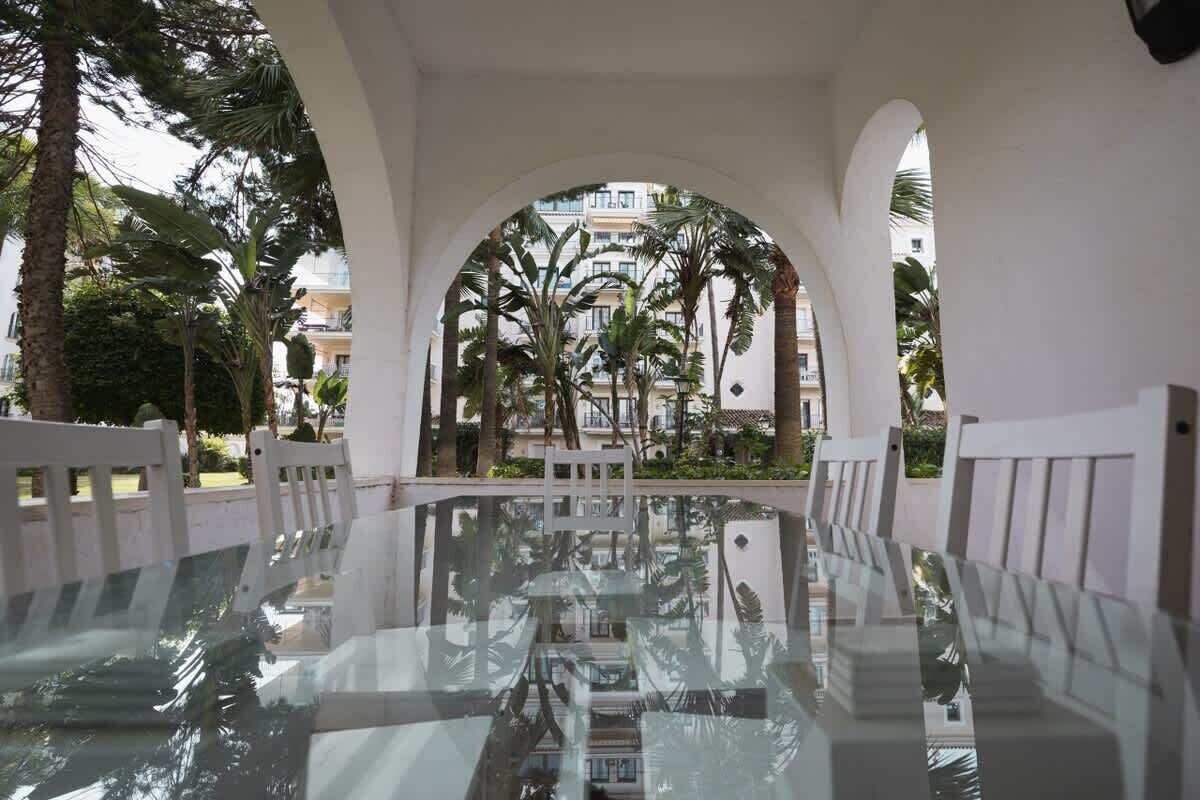 A glass-top table with chairs in a covered outdoor area, surrounded by white columns and palm trees.