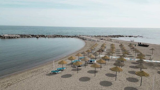 En strand med rækker af liggestole og parasoller, et stenmolle og roligt hav.