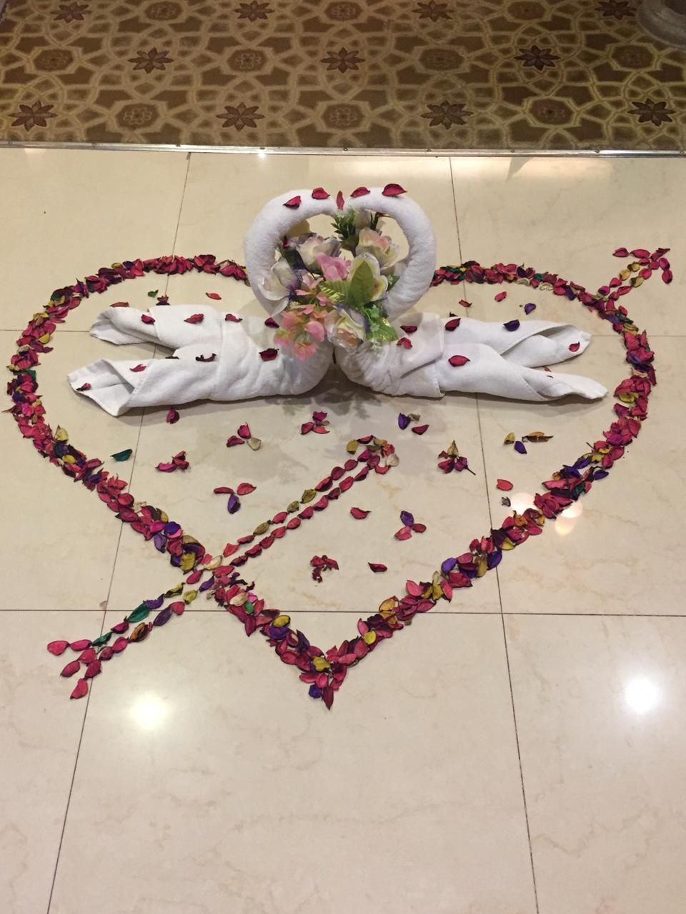 A decorative arrangement of white swans made from fabric, surrounded by a heart-shaped border of colorful flower petals on a tiled floor.