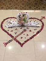 A decorative arrangement of white swans made from fabric, surrounded by a heart-shaped border of colorful flower petals on a tiled floor.