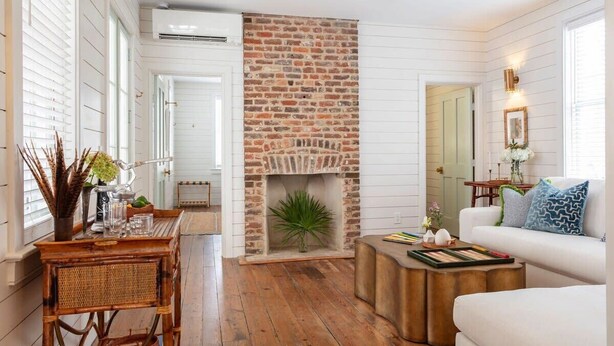 A living room with a brick fireplace, white walls, wooden flooring, a white sofa, and a coffee table. - Casa Zoë | Royal House – Charleston’s Crown (Charleston)