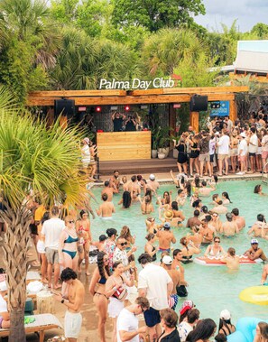 Un espace extérieur animé autour d’une piscine, avec une terrasse en bois et une scène pour des concerts.