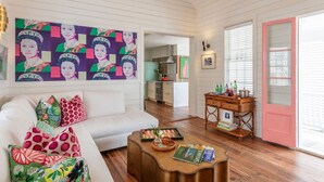 A living room with a white sofa, colorful pillows, a coffee table with books and a tray, a pink door, and a kitchen in the background. - Casa Zoë | Royal House – Charleston’s Crown (Charleston)