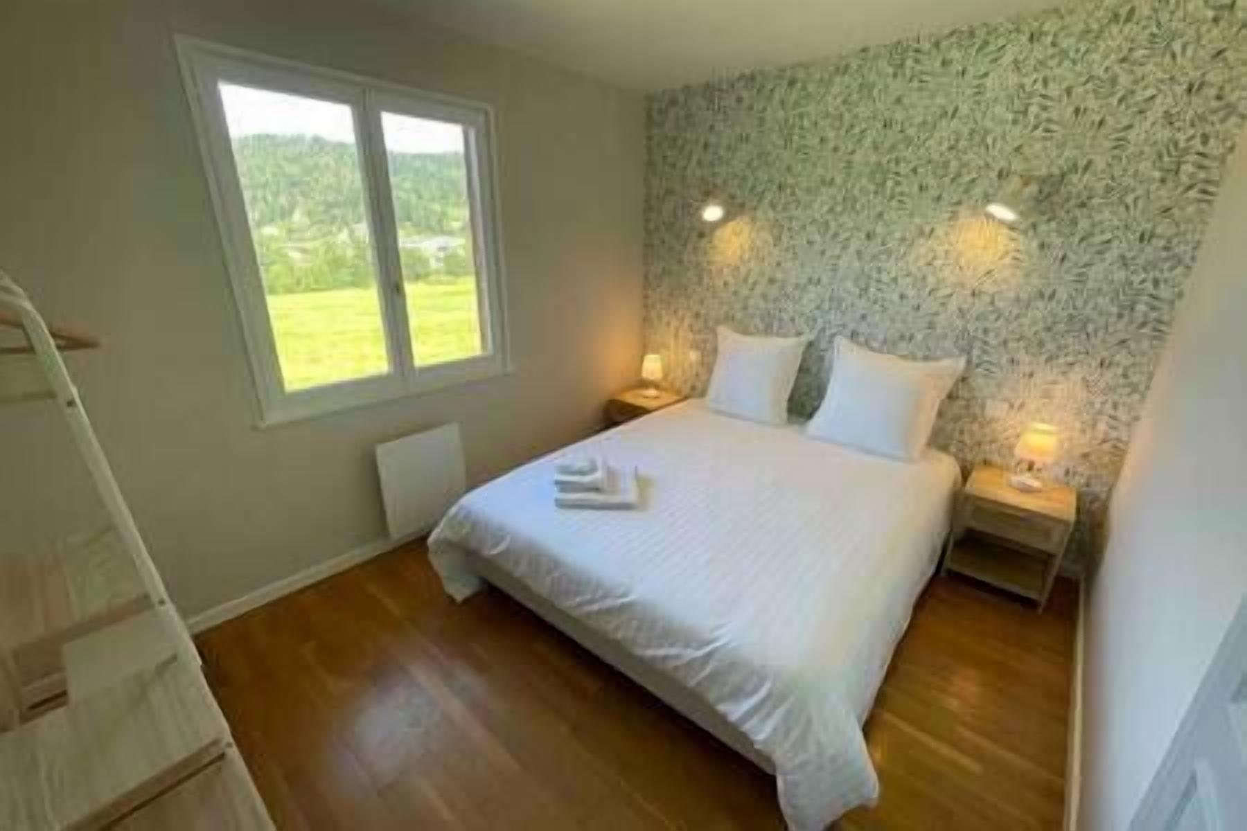 Quarto com uma cama grande, duas mesas de cabeceira com luminárias, uma janela com vista para o verde e piso de madeira.