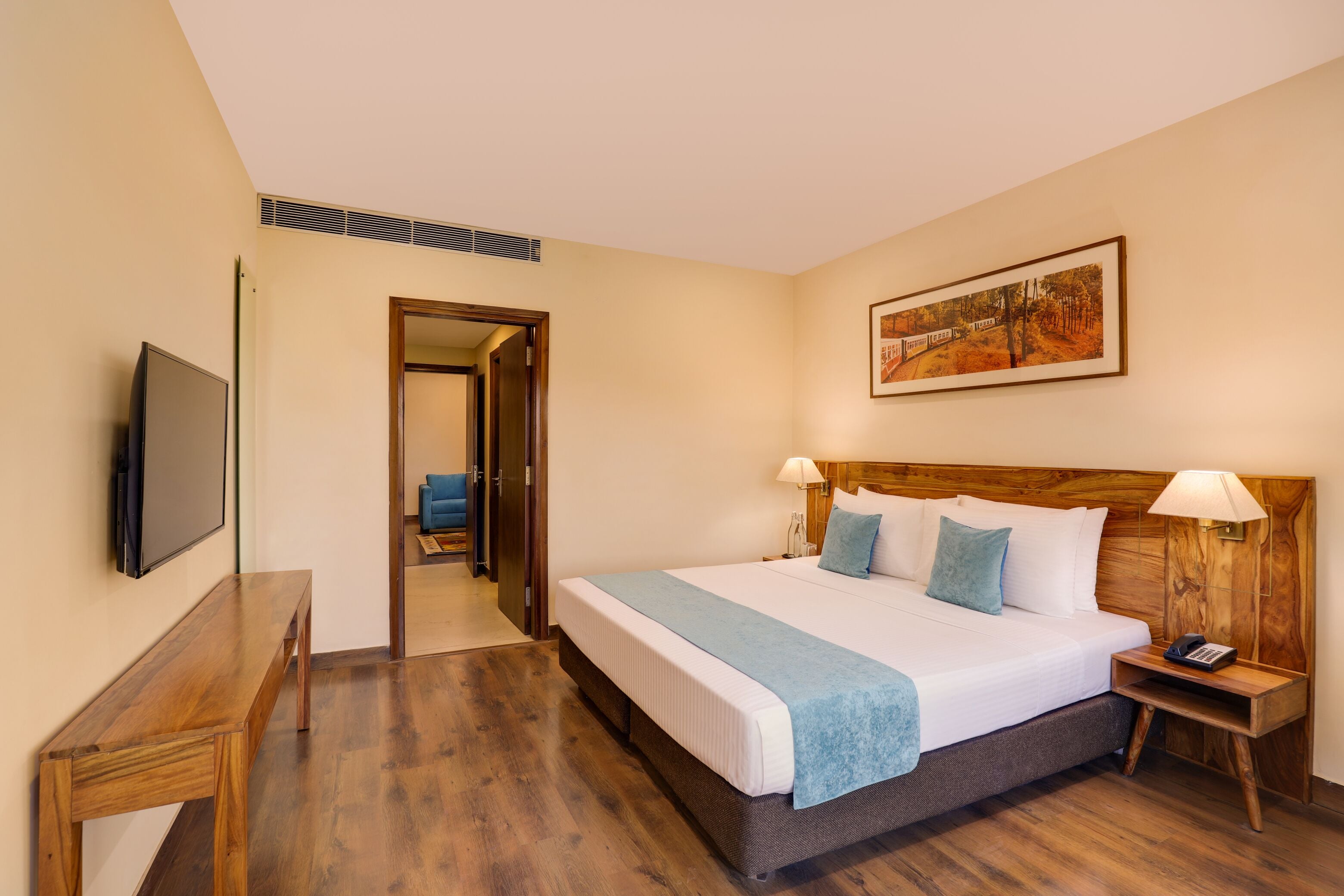 A hotel room with a large bed, wooden headboard, bedside lamps, a flat-screen TV, a wooden desk, and a framed picture on the wall.