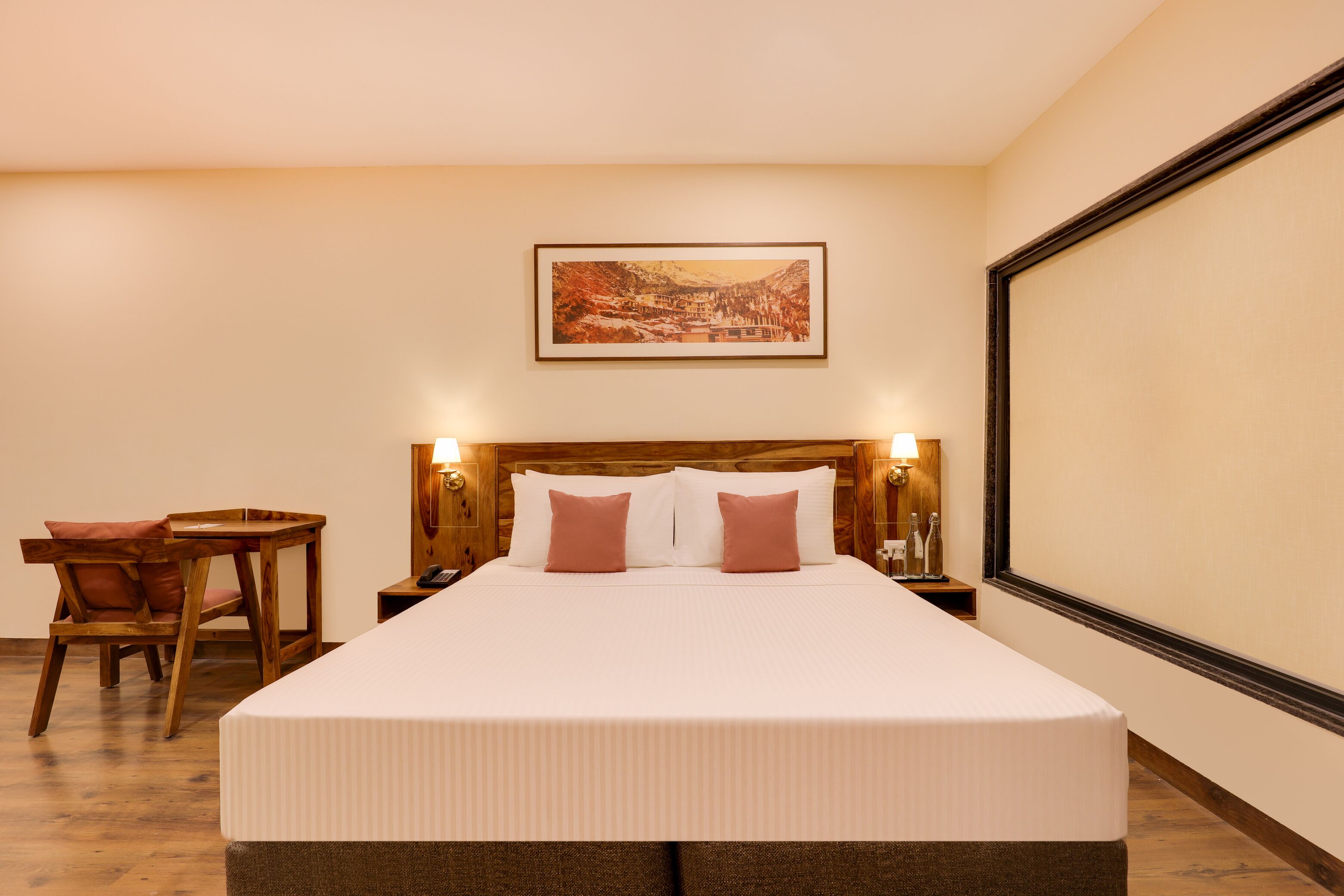 Habitación de hotel con una cama grande, cabecera de madera, un cuadro en la pared, una mesita con una silla y una mesita de noche con lámpara.