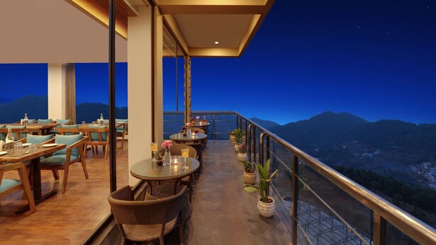 Un restaurant avec des tables et des chaises, une rambarde en verre et une vue sur les montagnes au crépuscule.