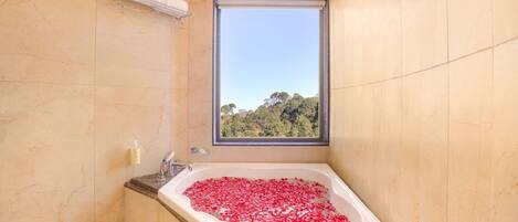Un baño con una bañera grande llena de pétalos de rosa, una ventana con vistas a árboles y un perchero para toallas.