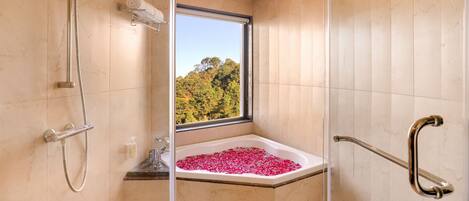 A modern bathroom with a shower, a bathtub filled with rose petals, and a window offering a view of trees.