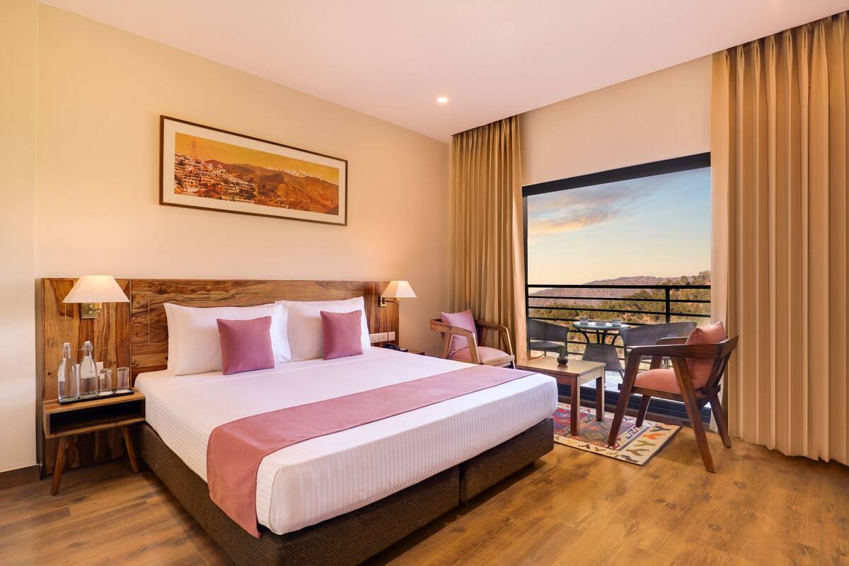 Habitación de hotel con una cama grande, cabecera de madera, dos mesitas de noche con lámparas, una pequeña mesa con sillas y un balcón con vistas.