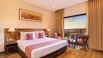 Habitación de hotel con una cama grande, cabecera de madera, dos mesitas de noche con lámparas, una pequeña mesa con sillas y un balcón con vistas.