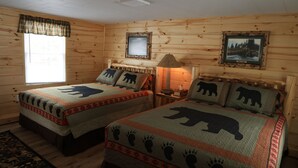 Ein blockhaussähnliches Zimmer mit zwei Betten, jeweils mit Bärenmotiv-Bettdecken, einem Fenster mit Karo-Vorhängen, einem gerahmten Bild an der Wand und einer Lampe auf einem Nachttisch.