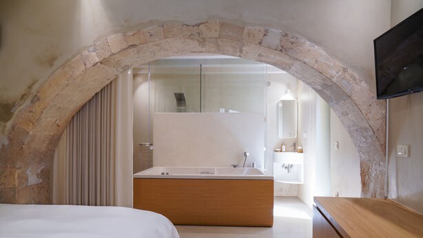 A bedroom with a large arched stone structure, a wooden bed, a white bathroom with a large mirror, and a flat-screen TV mounted on the wall.