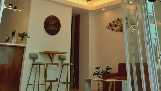 L’entrée d’un hôtel, avec un comptoir d’accueil, un espace salon et une enseigne indiquant « Hôtel Cinta ».