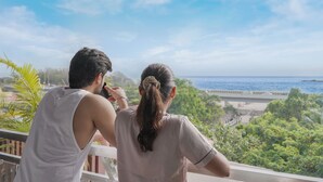 Deux personnes se tiennent sur un balcon donnant sur une plage, sous un ciel dégagé.