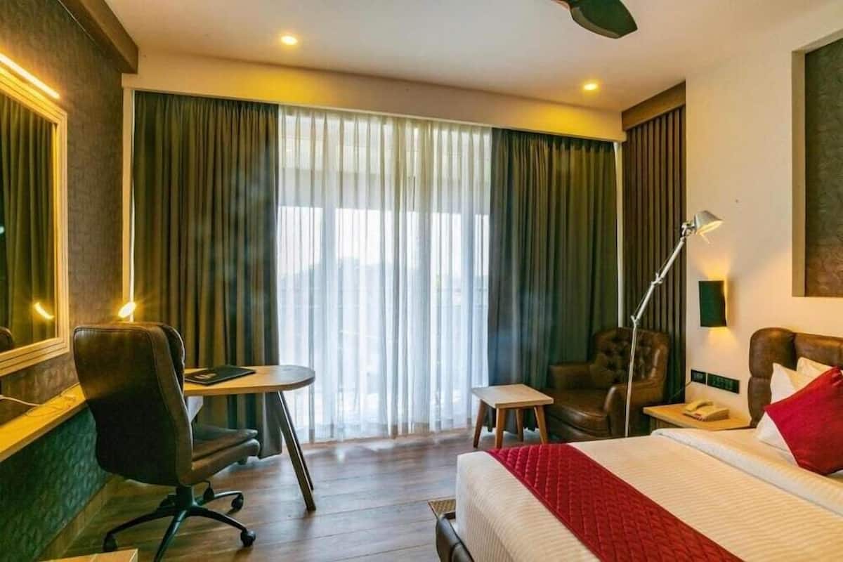 A hotel room with a bed, a desk, a chair, a lamp, and a large window with curtains.