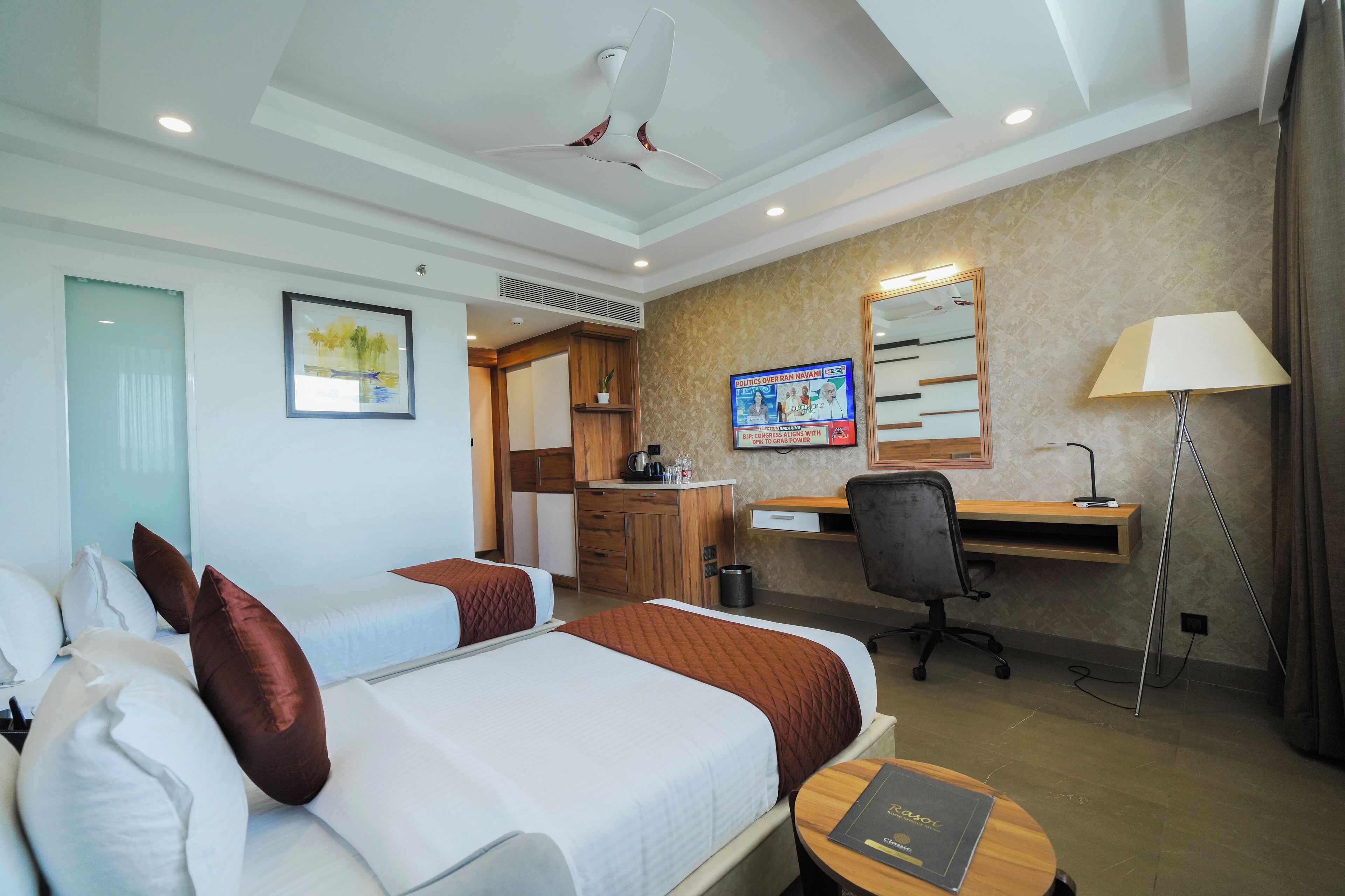 A hotel room with two beds, a desk, a chair, a lamp, and a TV.