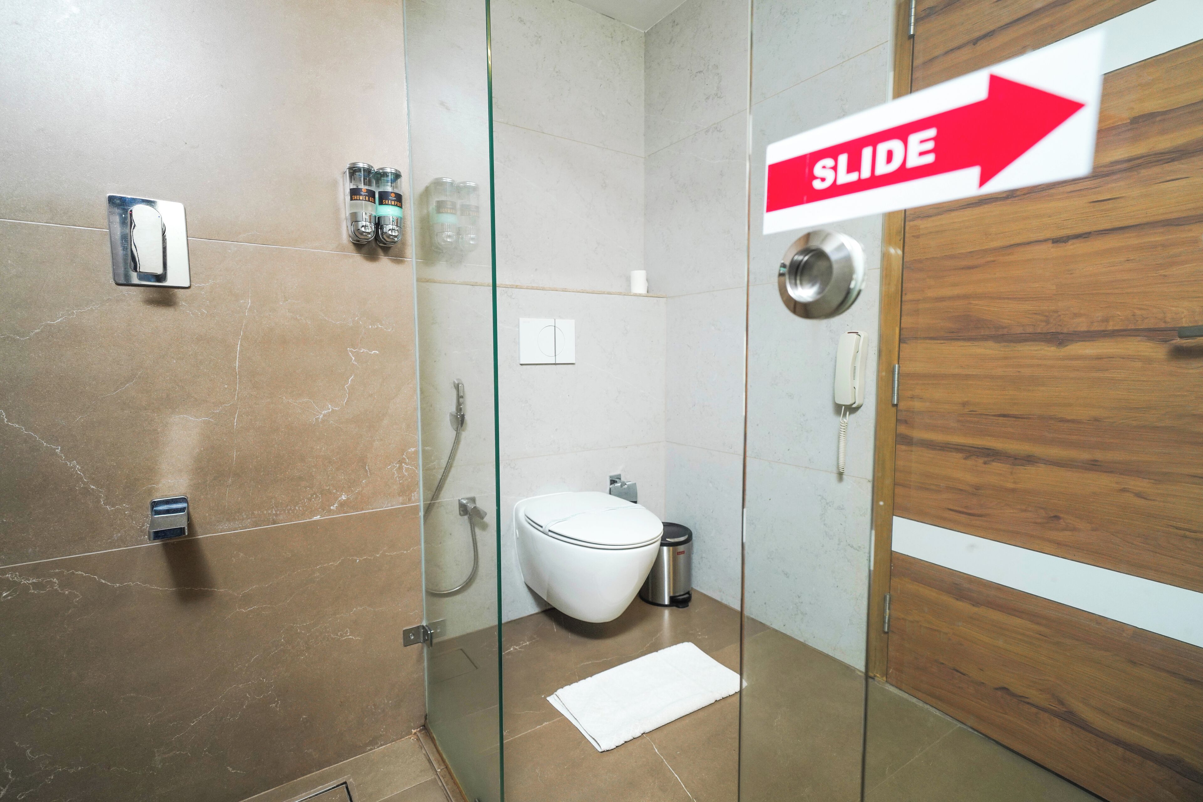A modern bathroom with a glass shower enclosure, a white toilet, and a wooden door with a 'SLIDE' sign.