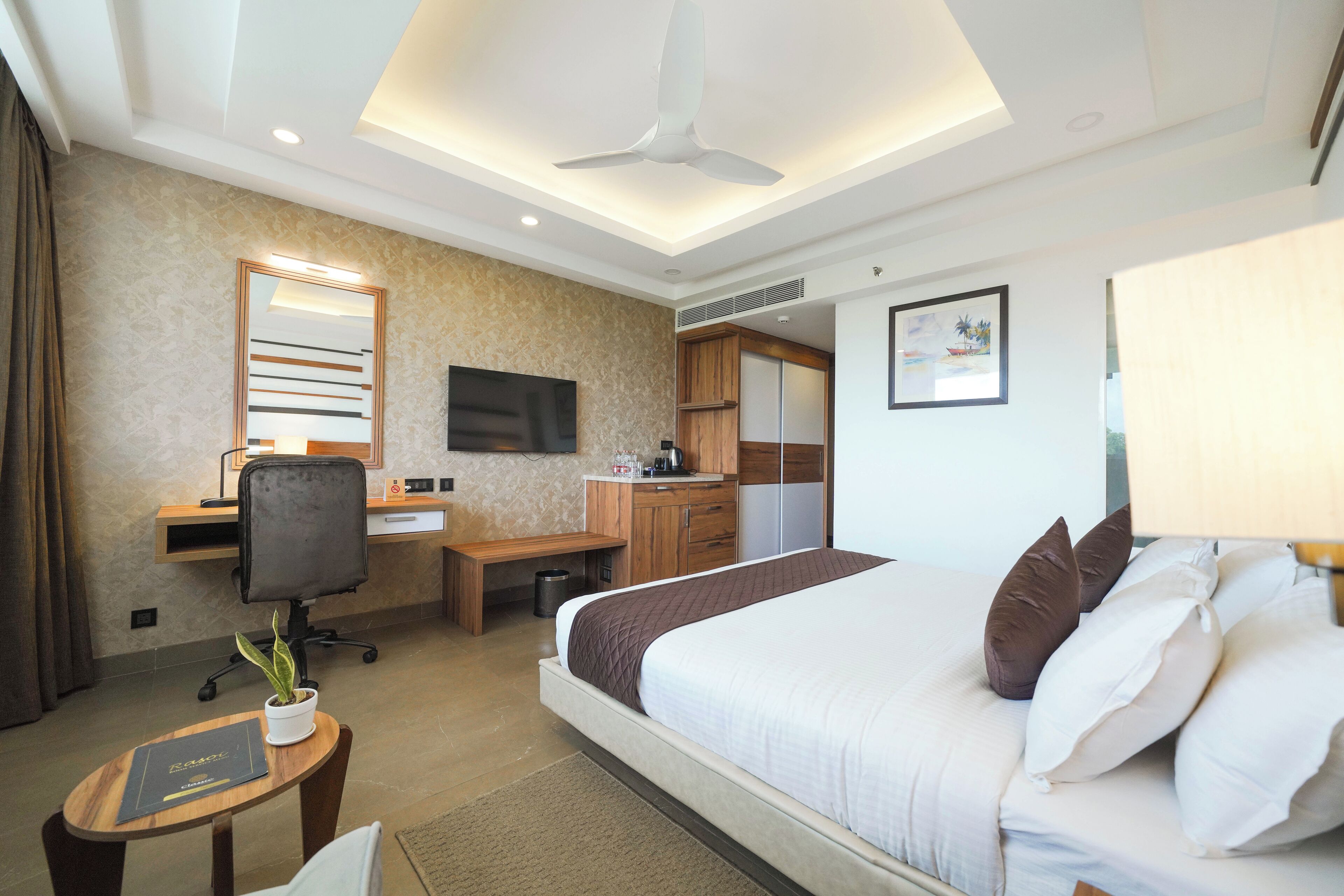 Habitación de hotel con una cama grande, un escritorio con computadora, una silla, una pequeña mesa con una planta y un ventilador de techo.