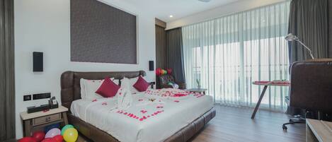 Una habitación de hotel moderna con una cama grande, un escritorio, una silla, un ventilador de techo y vistas al exterior a través de amplias ventanas.