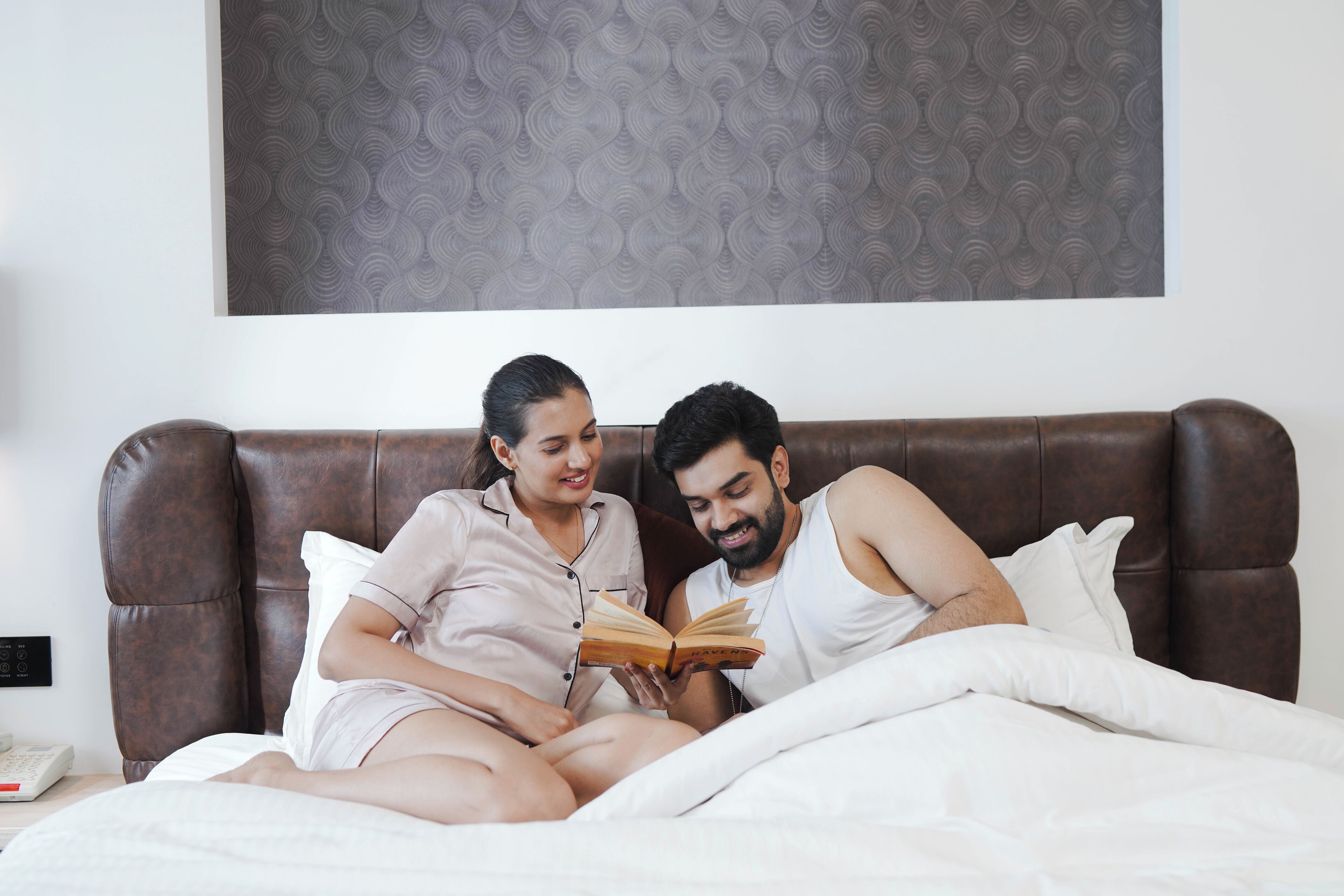 A couple sitting on a bed reading a book in a room with a patterned headboard and a plain wall.