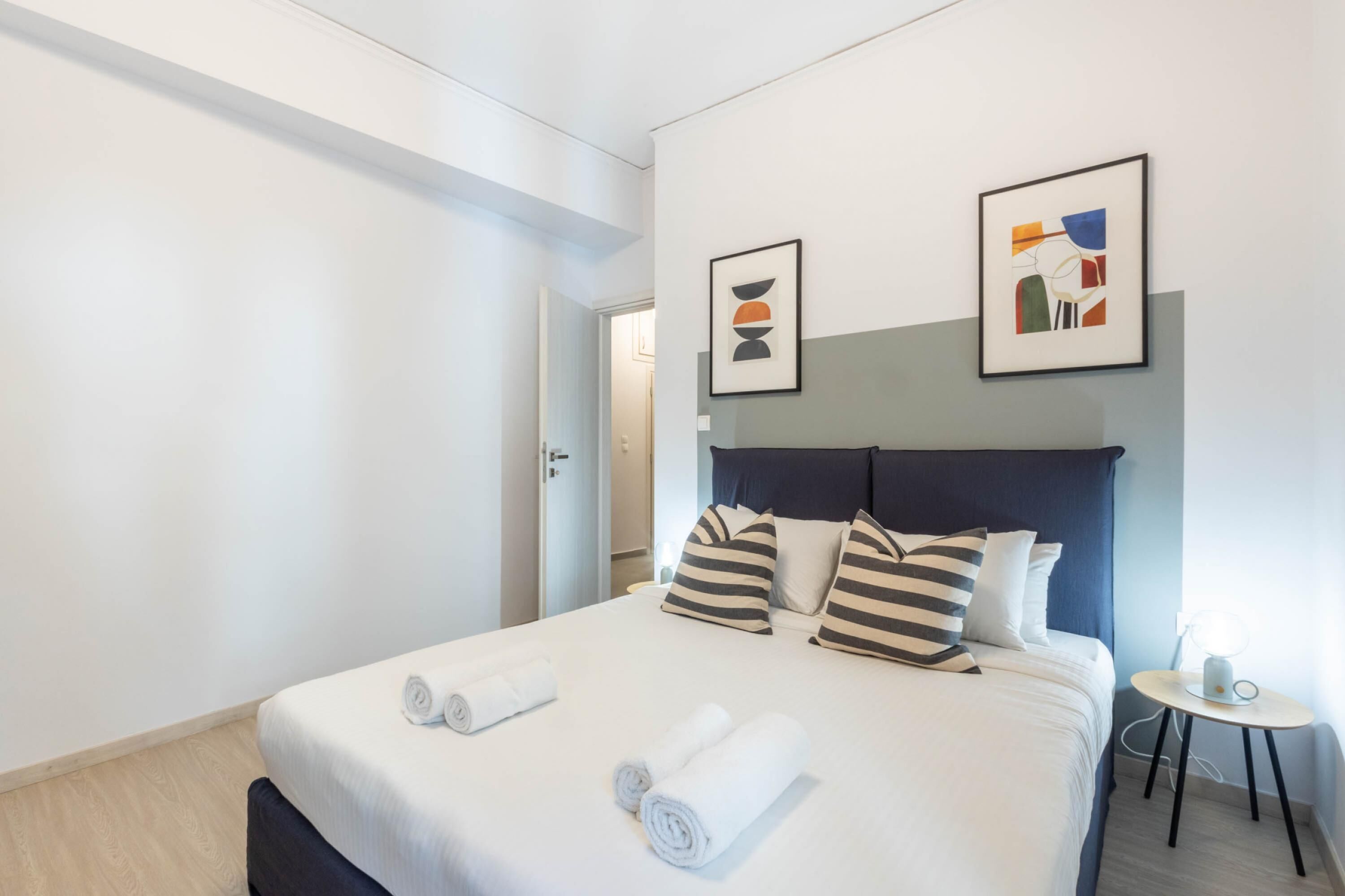 A hotel room with a neatly made bed, two pillows, a bedside table with a lamp, and two framed pictures on the wall.