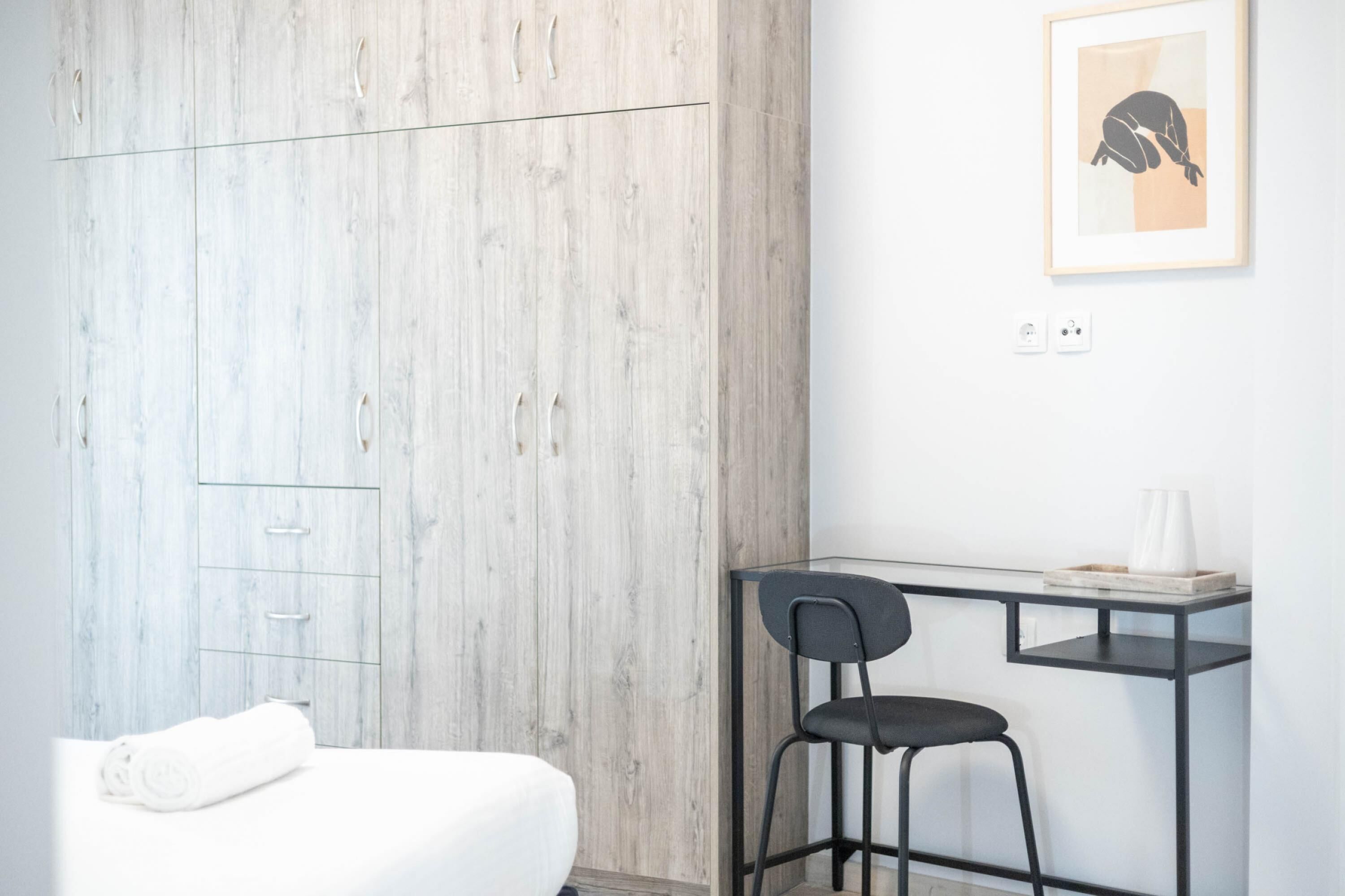 A minimalist room with a wooden wardrobe, a white bed, a black chair, and a small desk with a framed picture on the wall.