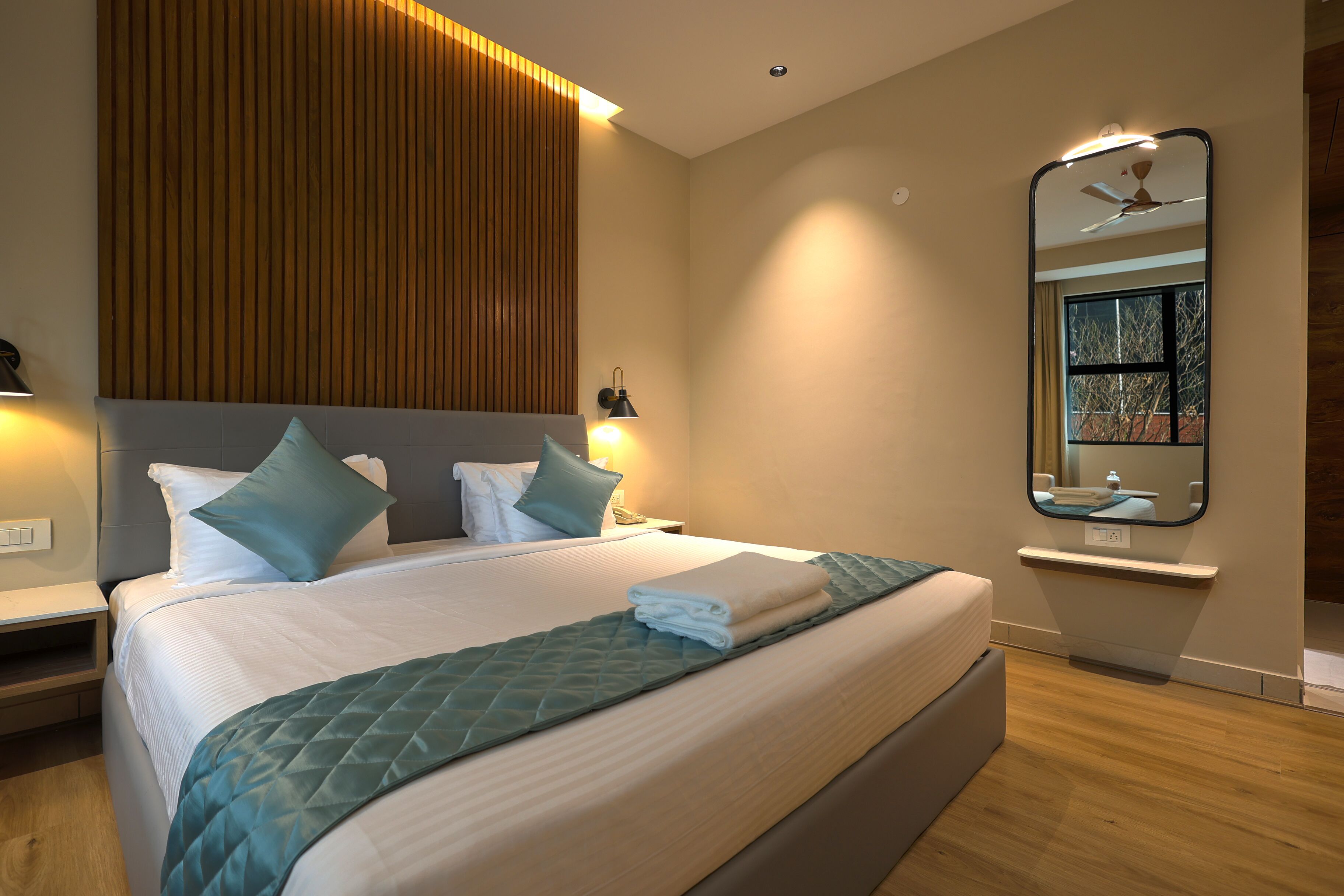 A modern hotel room with a large bed, a wooden panel wall, a nightstand, a mirror, and a window with a view of trees.
