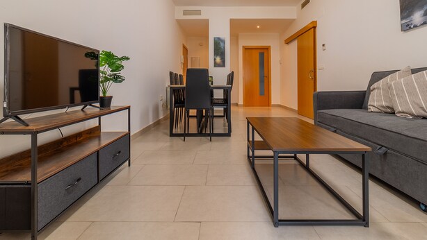 A modern living room with a flat-screen TV, a wooden coffee table, a grey sofa, and a dining area with a table and chairs.