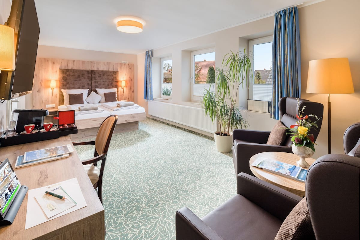 A hotel room with a bed, a desk with a laptop, a chair, a sofa, a table with a lamp, and a window with curtains.