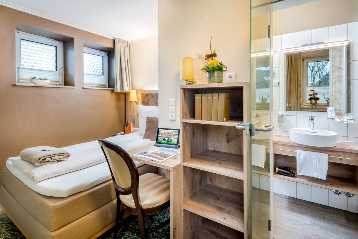 A hotel room with a bed, a desk with a laptop, a chair, a small shelf with books, a mirror, and a bathroom with a sink and a towel rack.