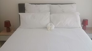 Una cama bien hecha con sábanas y almohadas blancas, dos mesitas de noche con lámparas y una pared lisa.