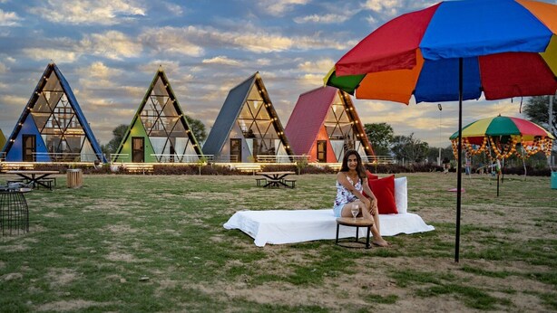 A woman sitting on a white lounge chair under a colorful umbrella in a grassy area with multiple triangular tents in the background. - Sunflower Saga (Jamwa Ramgarh)