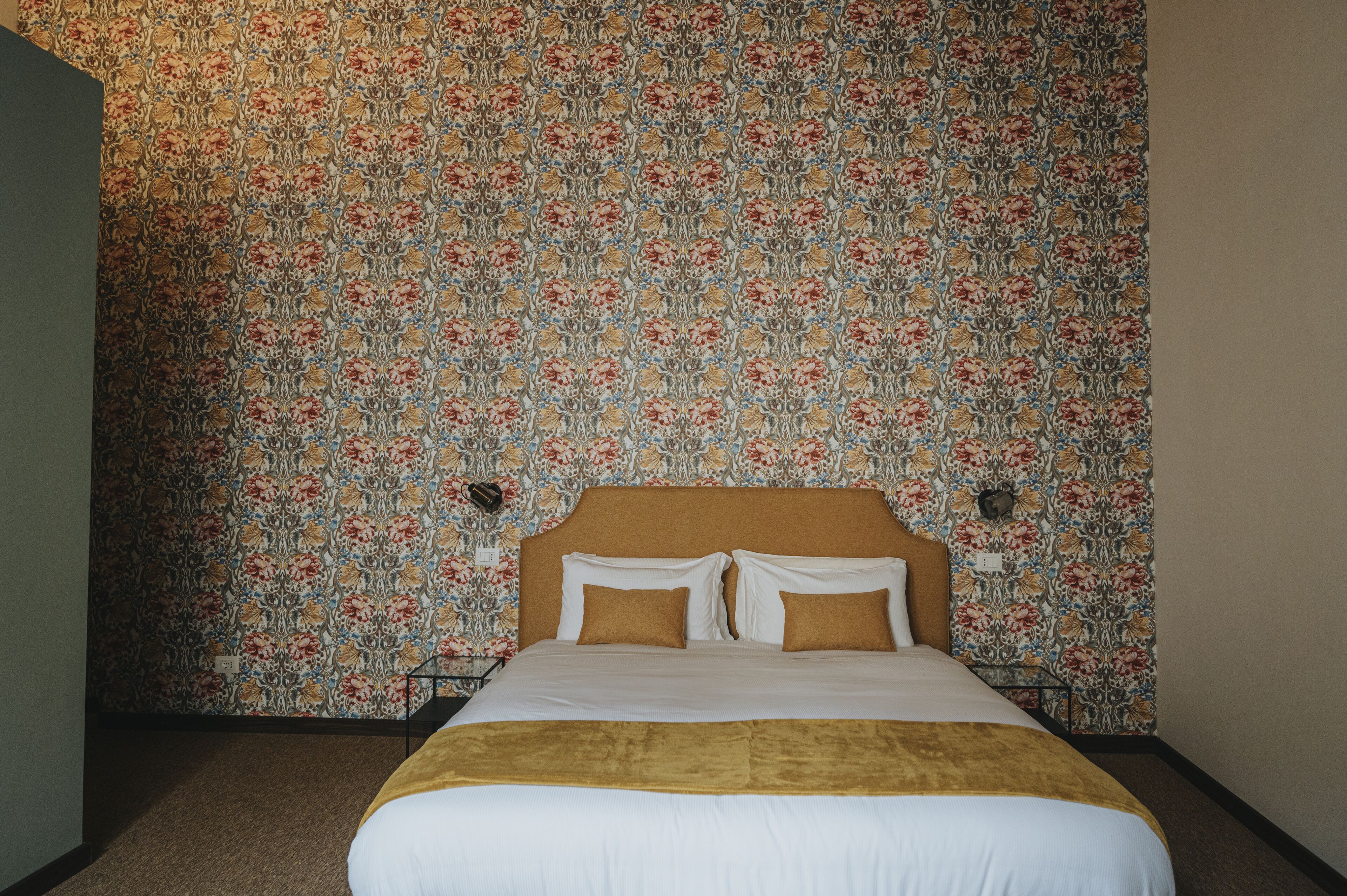A neatly made bed with a brown headboard, a floral-patterned wallpaper, and a brown throw blanket.