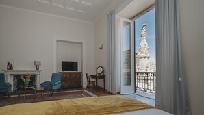 A hotel room with a bed, a TV, a fireplace, a desk, and a view of a historic building through a balcony. - Palazzo Pandola - Luxury Suites (Naples)